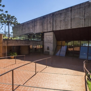 Fachada do Auditório Claudio Santoro. O céu está azul e à esquerda há algumas araucárias. A fachada o Auditório é composta por um prédio arquitetônico com cimento aparente. Há uma vidraça e também há uma escada larga com corrimão.