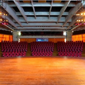 Na imagem há o palco do Auditório Claudio Santoro em primeiro plano e em segundo plano poltronas vermelhas divididas em cinco setores. Acima há estrutura arquitetônica do Auditório com recortes da laje. E aos fundos do espaço, há cortinas laranjas.