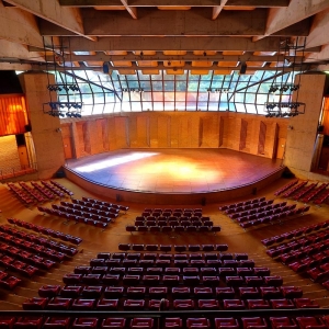 A imagem mostra o Auditório Claudio Santoro que possui uma arquitetura que se assemelha a arena. Há 814 poltronas na cor vermelha, um palco com luzes e vidraças nas quais é possível observar a natureza.