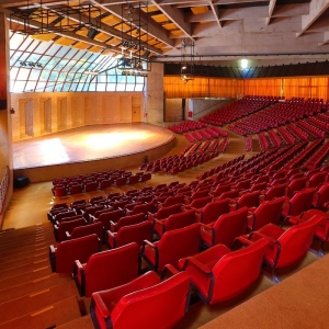 Na imagem há o palco do Auditório Claudio Santoro em primeiro plano e em segundo plano poltronas vermelhas divididas em cinco setores. Acima há estrutura arquitetônica do Auditório com recortes da laje. E aos fundos do espaço, há cortinas laranjas.
