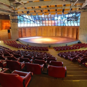 A imagem mostra o Auditório Claudio Santoro que possui uma arquitetura que se assemelha a arena. Há diversas poltronas na cor vermelha, um palco com luzes e vidraças nas quais é possível observar a natureza.