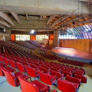 A imagem mostra o Auditório Claudio Santoro que possui uma arquitetura que se assemelha a arena. Há diversas poltronas na cor vermelha, um palco com luzes e vidraças nas quais é possível observar a natureza.