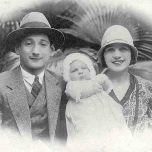 A imagem está em preto e branco e mostra Felícia Leirner bebê sendo segurada no colo pelos pais. Há um homem, jovem, de terno e chapéu. Um bebe de vestido e touca. Uma mulher, jovem também de chapéu.