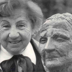 A imagem está em preto e branco e mostra Felícia Leirner idosa ao lado de uma escultura. Felícia tem cabelos cacheados, algumas marcas de expressões, olhos claros. Ela usa aparentemente uma camisa e um casaco. Ao seu lado há uma escultura que aparenta ser o rosto de uma mulher.