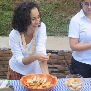 Uma mulher branca, de cabelos pretos com alguns fios grisalhos está segurando um biscoito nas mãos. A sua frente a uma mesa com doces e biscoitos.