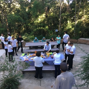 Área de piquenique em meio a natureza, com mesas e bancos. Na mesa há diferentes tipos de comidas como pão, presunto, biscoito, queijo, pão, etc. Ao redor da mesa há pessoas se servindo.