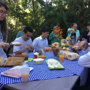 Mesa com diferentes tipos de comidas como pão, presunto, biscoito, queijo, pão, etc. Ao redor da mesa há pessoas se servindo.