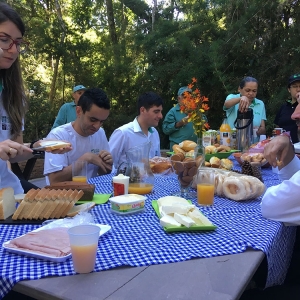 Mesa com diferentes tipos de comidas como pão, presunto, biscoito, queijo, pão, etc. Ao redor da mesa há pessoas se servindo.