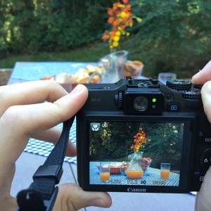 Uma câmera enquadrando a imagem de uma mesa com uma jarra, copos com suco, e um jarro de flores.