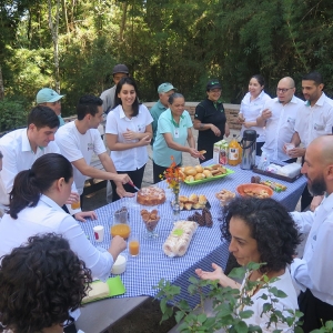 Ao redor de uma mesa com diferentes tipos de comidas como pão, presunto, biscoito, queijo, pão, etc. há pessoas se servindo durante o piquenique.