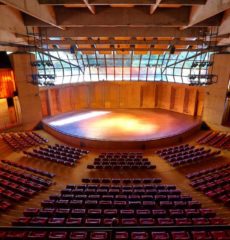 A imagem mostra o Auditório Claudio Santoro que possui uma arquitetura que se assemelha a arena. Há diversas poltronas na cor vermelha, um palco com luzes e vidraças nas quais é possível observar a natureza.