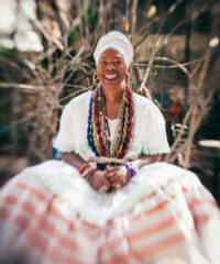 Nega Duda, uma mulher negra que usa um turbante branco, colares e um vestido longo branco