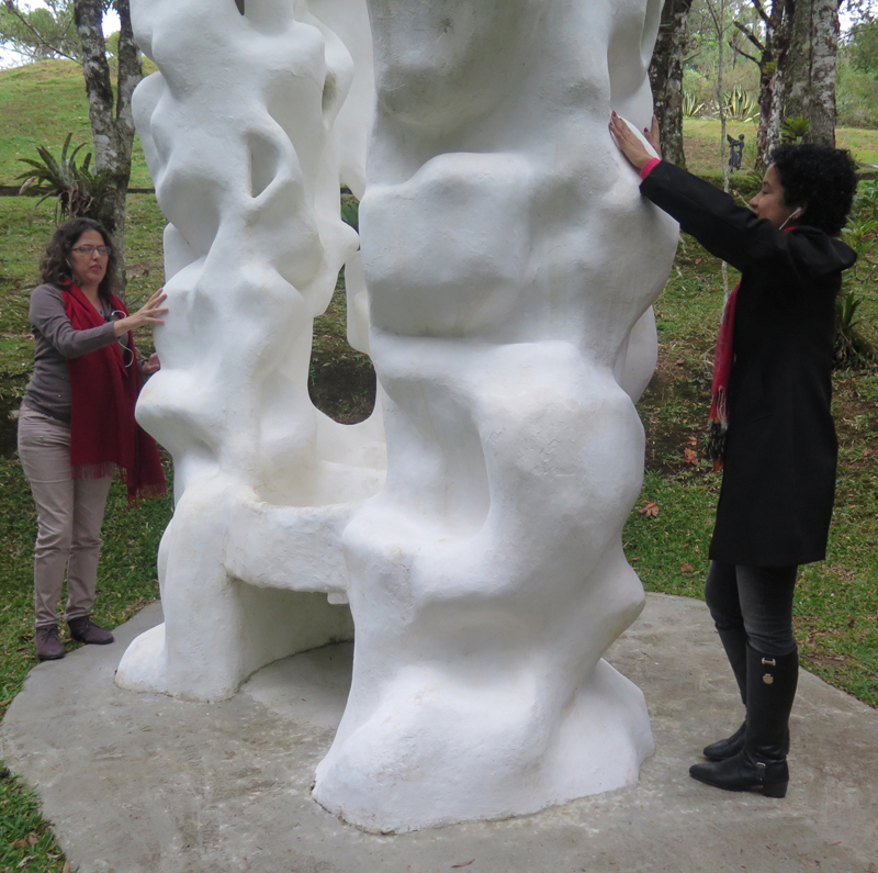 Duas mulheres, sendo uma branca e outra negra, tocam a obra "Três colunas" de Felícia Leirner sentido os contornos da obra por meio do tato.