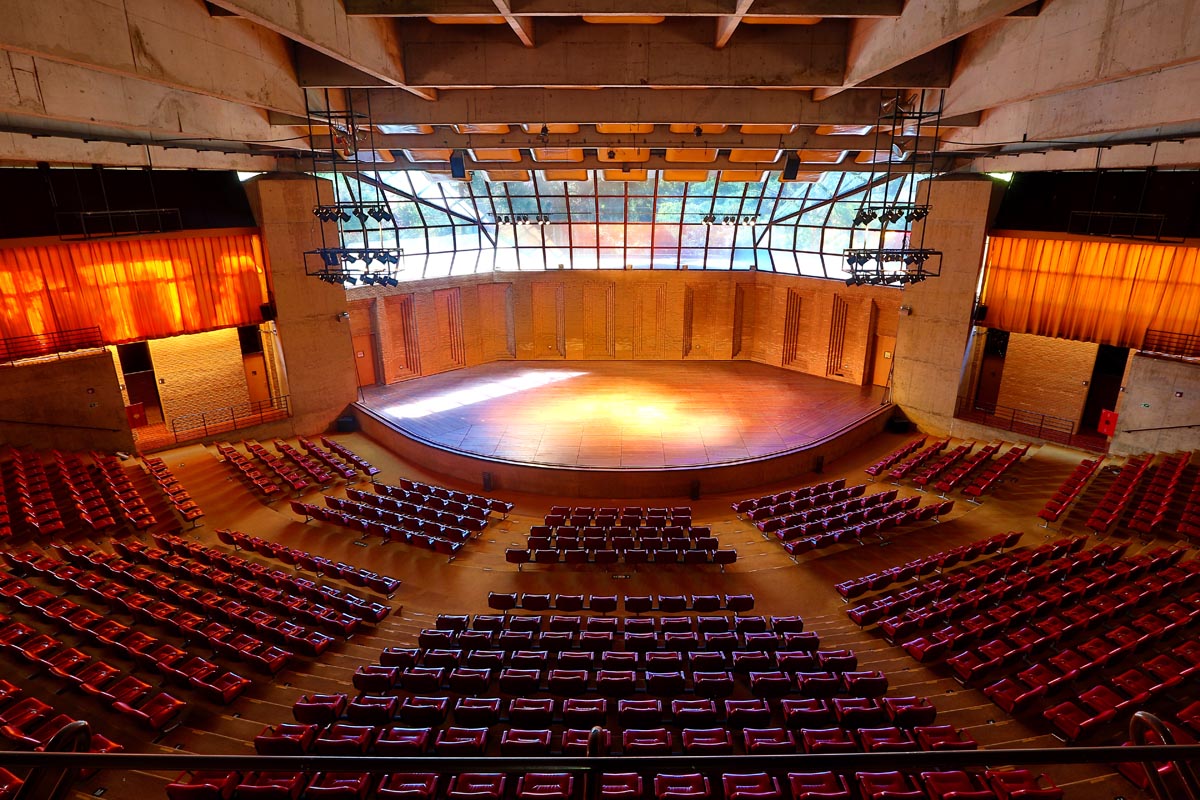 A imagem mostra o Auditório Claudio Santoro que possui uma arquitetura que se assemelha a arena. Há diversas poltronas na cor vermelha, um palco com luzes e vidraças nas quais é possível observar a natureza.