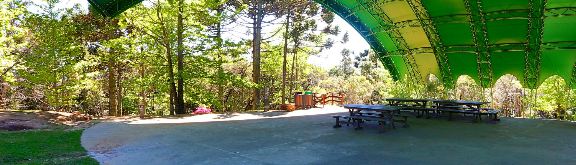 Concha acústico do Museu Felícia Leirner. É um espaço amplo com uma cobertura em lona verde. Há algumas mesas e bancos no centro do espaço. Ao ar livre, a vegetação ao redor compõe a paisagem