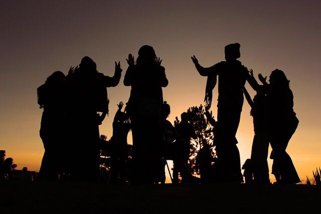 A imagem mostra o contorno de pessoas numa roda de dança sobre o por do sol