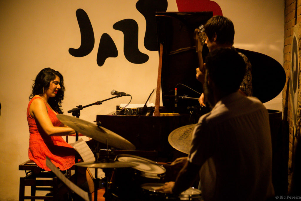 Uma mulher branca, de cabelos pretos e longos está sentada de frente para um piano, tocando o instrumento. Próximo a ela há outros dois músicos que tocam contrabaixo e bateria.