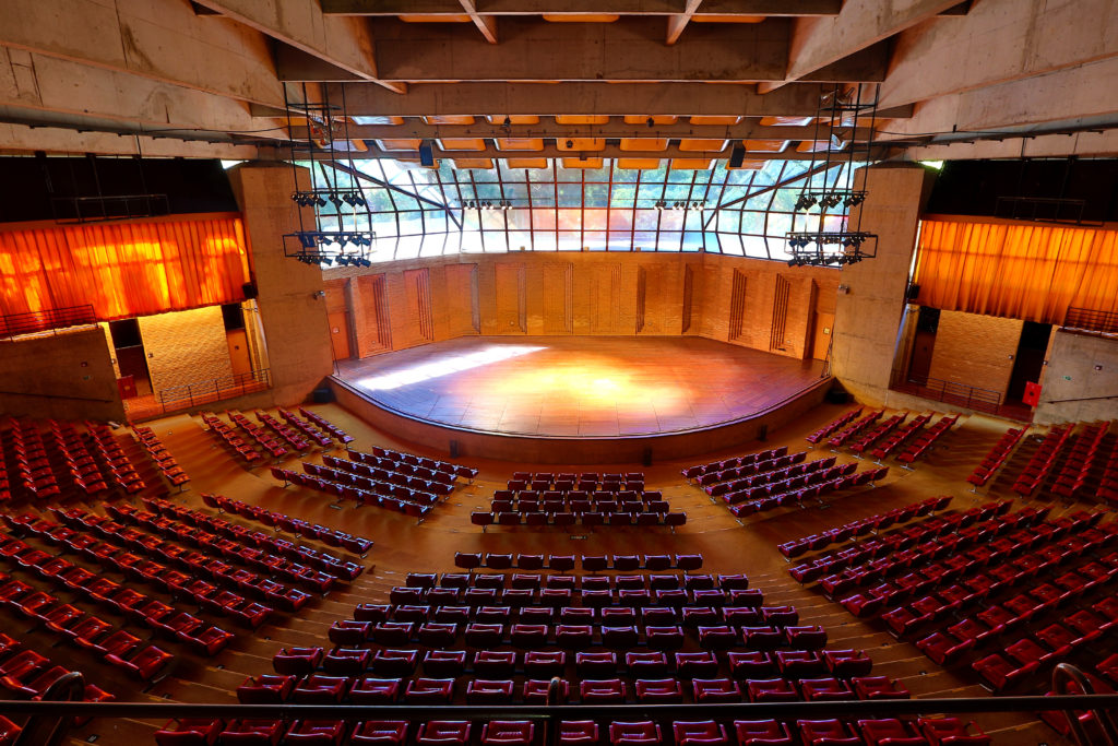 A imagem mostra o Auditório Claudio Santoro que possui uma arquitetura que se assemelha a arena. Há diversas poltronas na cor vermelha, um palco com luzes e vidraças nas quais é possível observar a natureza.