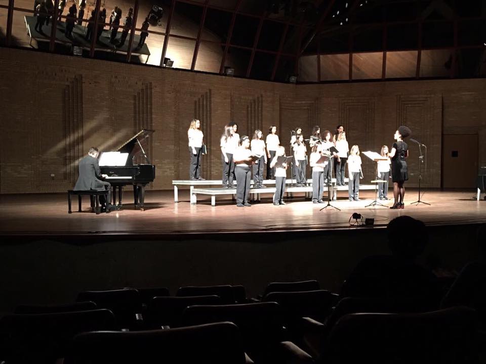 A imagem mostra um grupo de meninas no palco do Auditório Claudio Santoro. Elas cantam sob a regência de uma mulher alta. Um homem toca piano à esquerda.