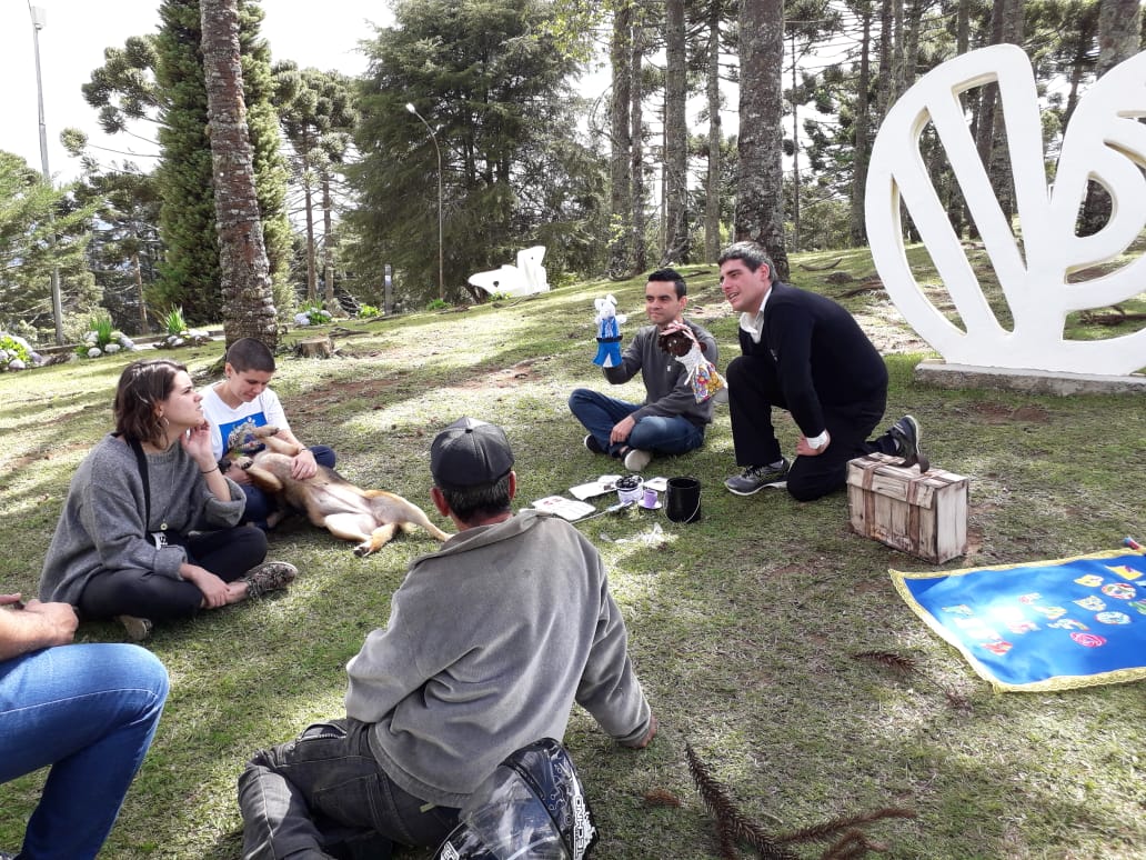 Pequeno grupo de pessoas está sentado na grama próximo a uma escultura. Dois educadores estão com bonecos em suas mãos e contam uma história ao grupo.