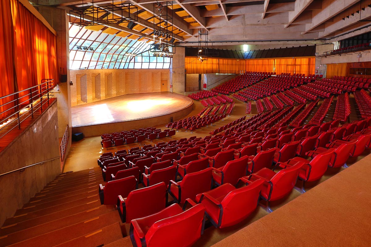 A imagem mostra o Auditório Claudio Santoro que possui uma arquitetura que se assemelha a arena. Há diversas poltronas na cor vermelha, um palco com luzes e vidraças nas quais é possível observar a natureza.