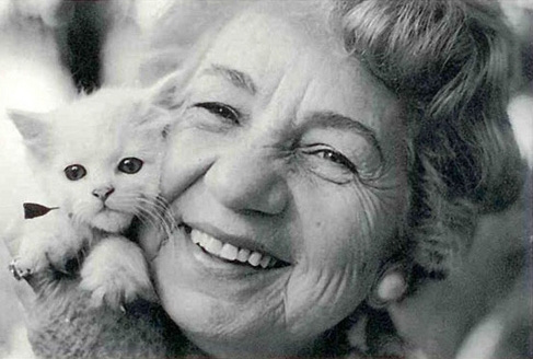 A imagem está preto e branco e há uma mulher idosa de cabelos curto e cacheados. Ela segura próximo ao seu rosto um filhote de gato.