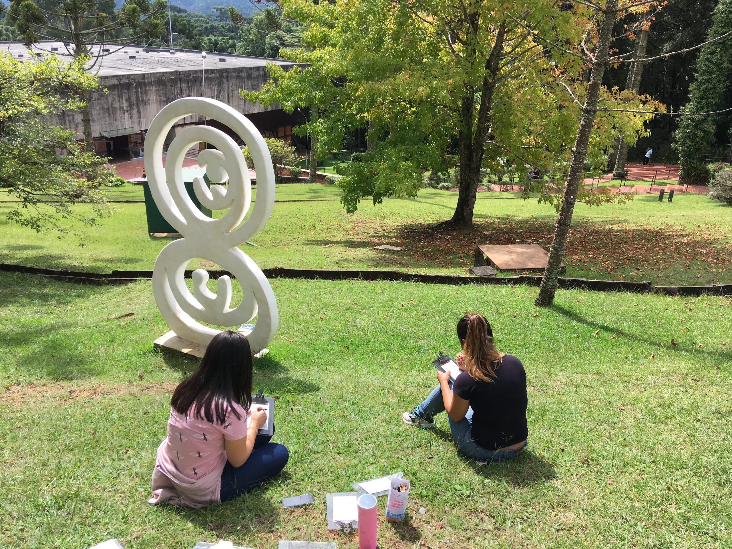 Duas mulheres estão sentadas próxima a escultura "Nascimento" e estão realizando uma atividade com uso de prancheta e lápis coloridos.