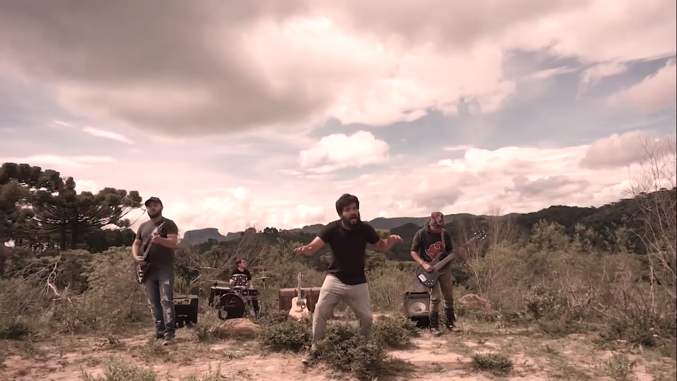 A imagem mostra uma manda composta por quatro homens que tocam seus instrumentos a céu aberto. A paisagem é composta por uma cadeia de montanhas na qual é possível ver a pedra do baú. Os músicos estão bem em meio a vegetação. Há esquerda, na frente, há um homem tocando guitarra. Ao fundo um homem toca bateria. Ao centro há um homem que está cantando e performando. À direita há outro homem tocando contra-baixo.