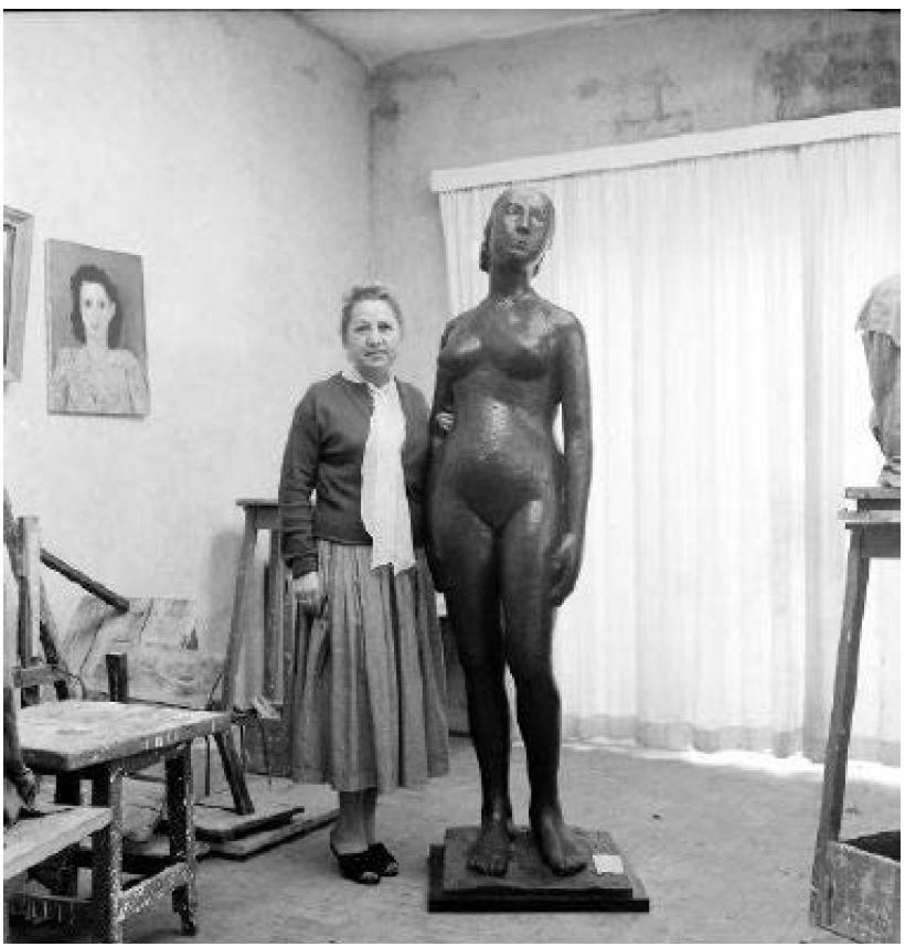 A imagem mostra Felícia Leirner em um ambiente que se assemelha a uma oficina ou ateliê. Ela está em pé ao lado esquerdo de uma escultura feita em bronze que representa uma mulher alta e com curvaturas.