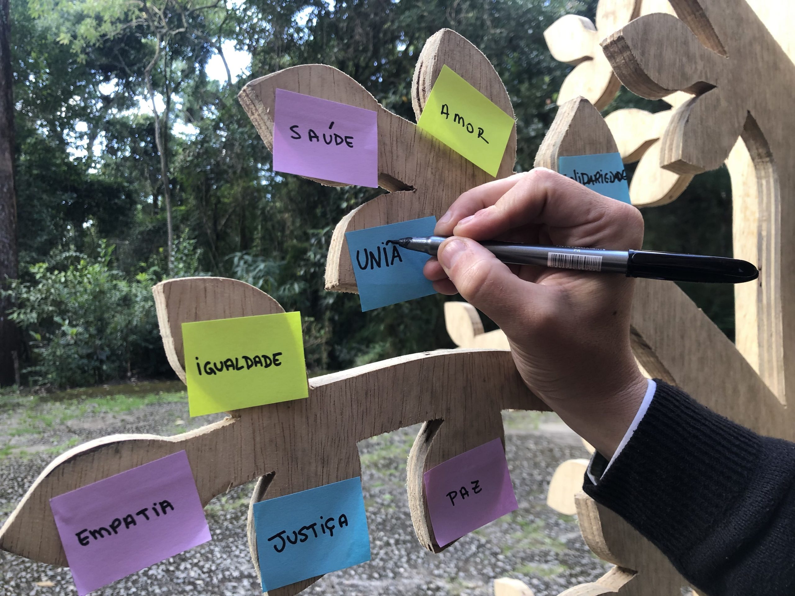 Na imagem, há galhos de uma árvore feita de madeira onde estão colados diversos post-it com palavras como: igualdade, empatia, saúde, amor. Uma mão está segurando uma caneta e escreve em um dos post it a palavra união.