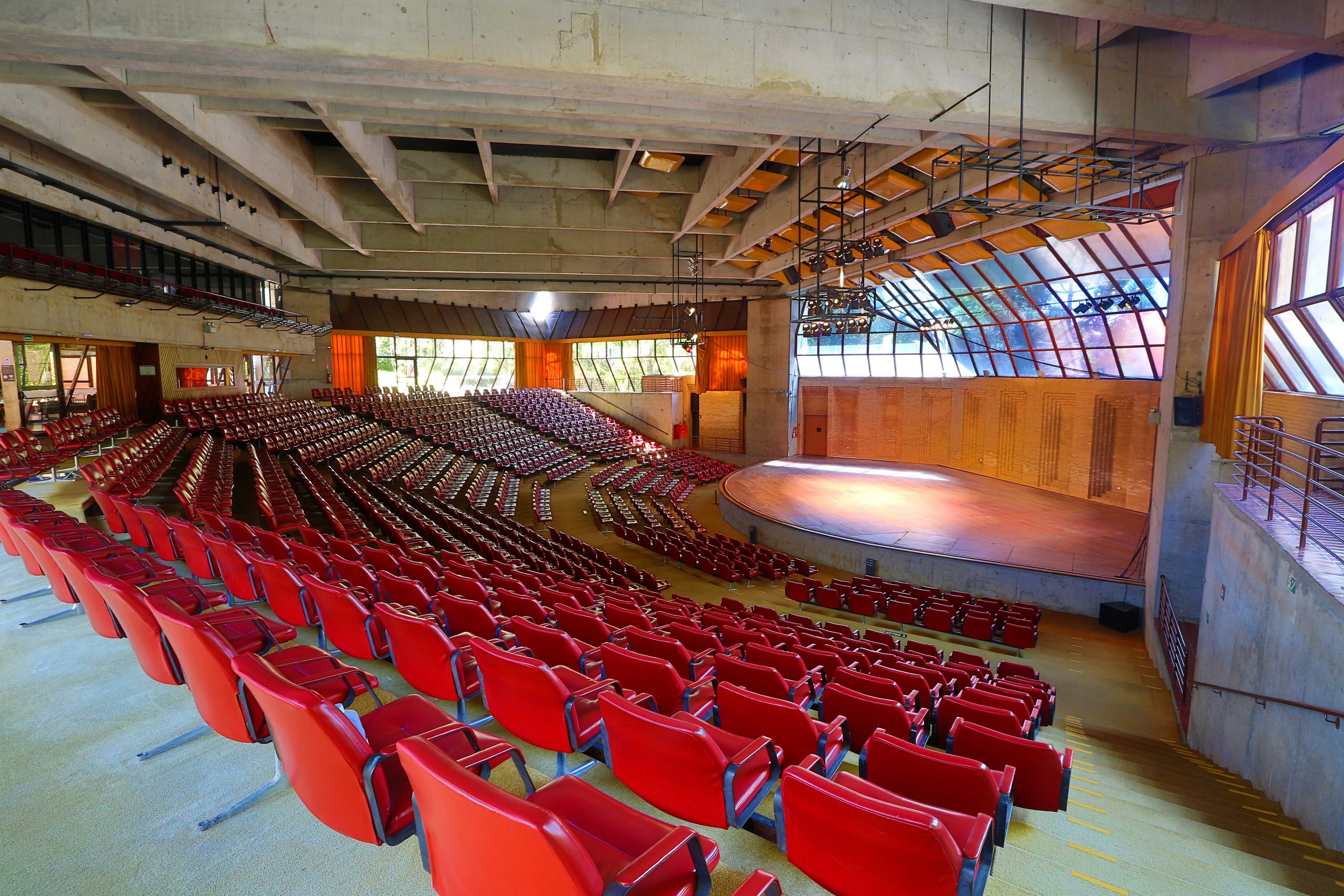 A imagem mostra o Auditório Claudio Santoro que possui uma arquitetura que se assemelha a arena. Há diversas poltronas na cor vermelha, um palco com luzes e vidraças nas quais é possível observar a natureza.