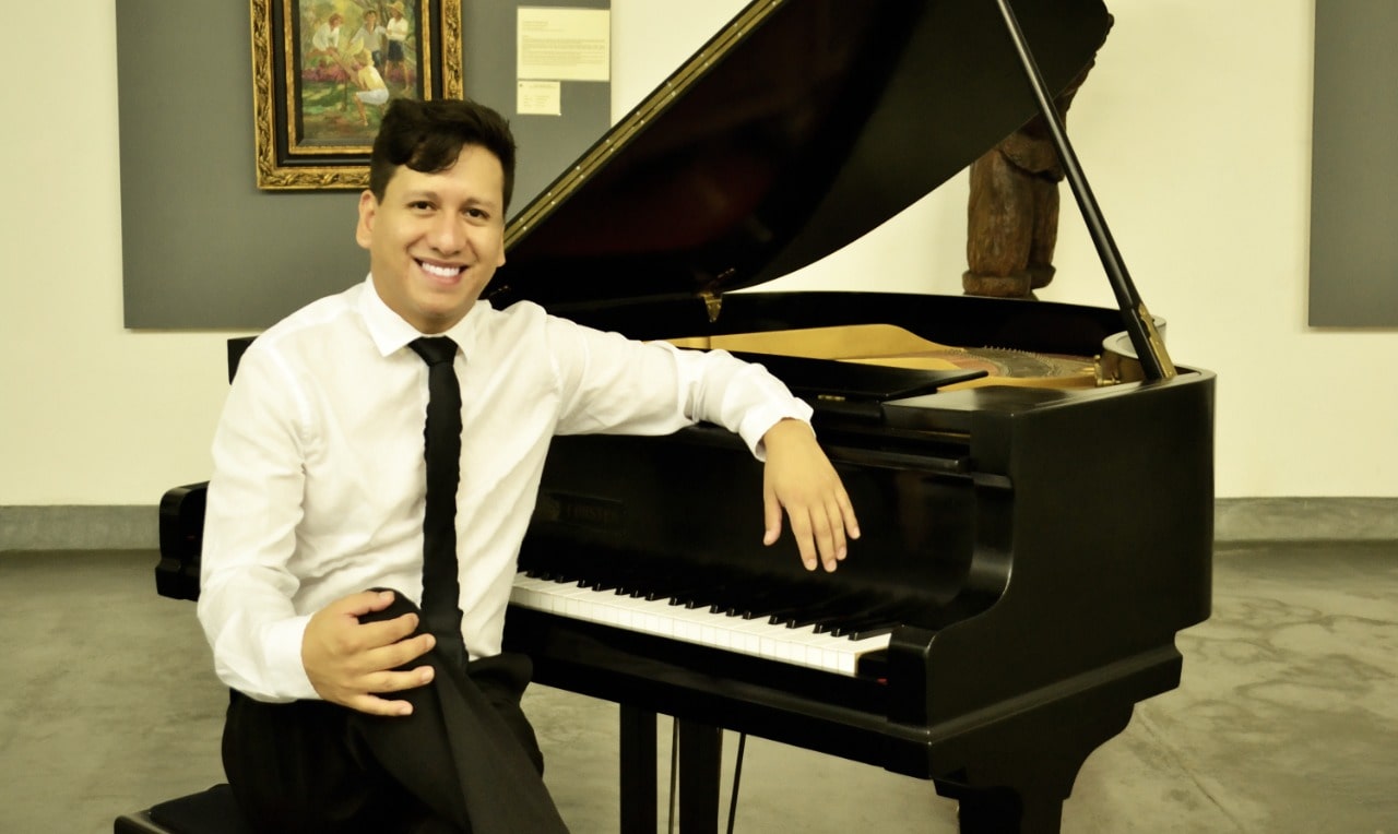 Um homem branco, de cabelos castanhos está usando uma camisa branca e uma gravata preta. Ele está sentado de frente com um piano de calda, apoiando um dos braços sobre o instrumento.