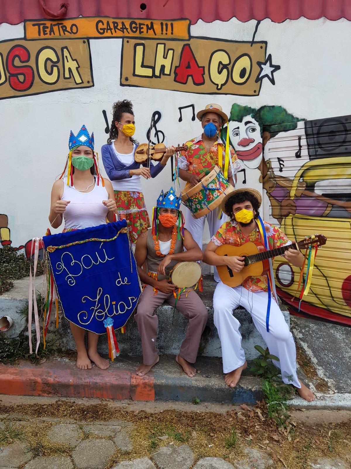 Cinco pessoas, brancas e negras, estão vestidas com peças de roupas brancas e floridas. Eles seguram em mãos instrumentos musicais de corda e percussão. Uma dessas pessoas, uma mulher branca, segura uma bandeira escrita "Baú de Folias".