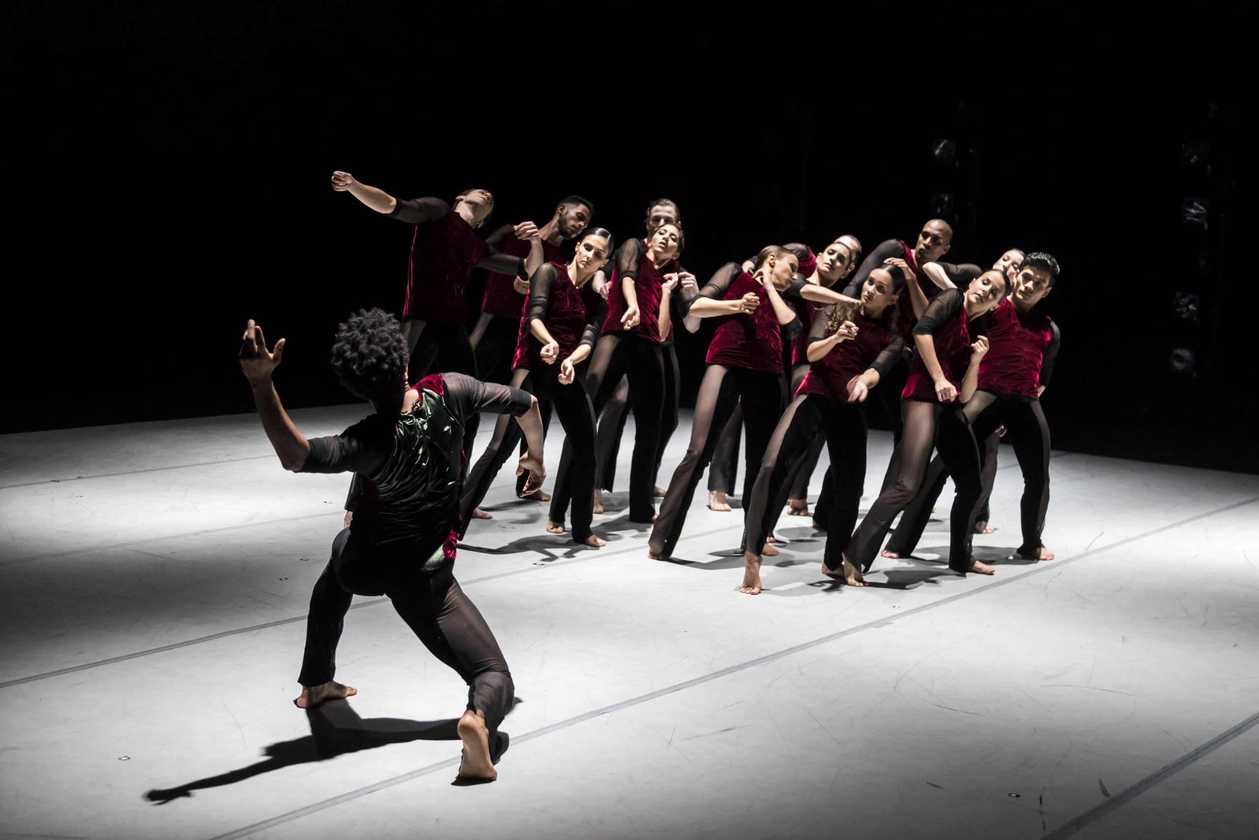 A imagem mostra um grupo de bailarinos dançando sobre um palco. Eles usam uma camiseta vermelha e uma calça preta.