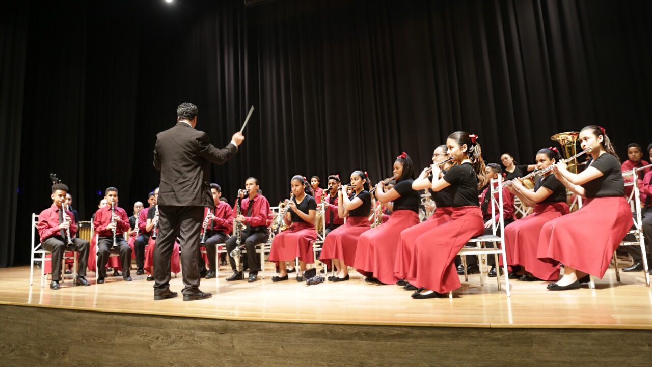 Crianças e adolescentes sentados em um palco tocam diferentes tipos de instrumentos, compondo uma orquestra. No centro, há uma homem de preto que faz a regência das crianças.