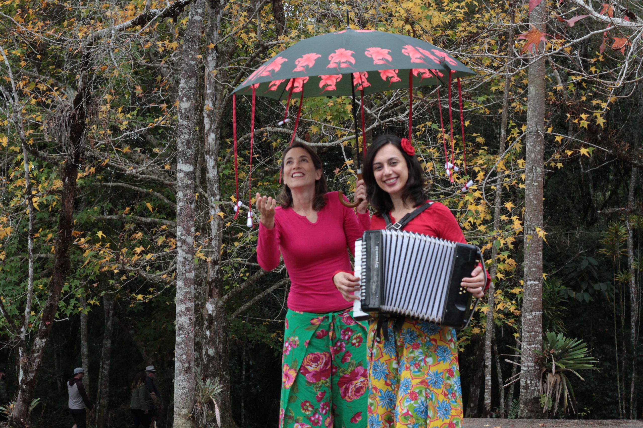 Na imagem há duas mulheres brancas ao ar livre tendo árvores e mata ao redor. Elas usam camisetas rosa e saias floridas. A mulher da esquerda segura num guarda chuvas e a mulher da direita está tocando acordeon.