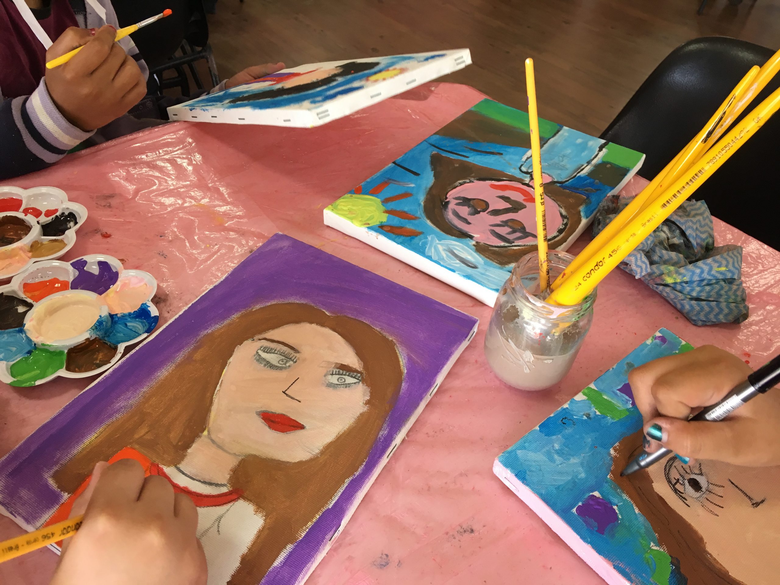 Sobre uma mesa há quatro quadros com pintura de autorretratos realizados por crianças.