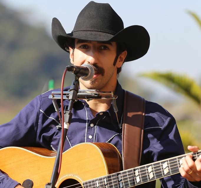 Na imagem, um homem branco usando um chapéu de cowboy na cor preta. Ele está tocando violão e cantando em frente a um microfone.