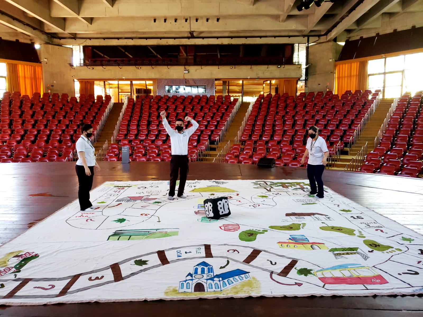 Na imagem há um jogo de tabuleiro gigante, feito de tecido, sobre o palco do Auditório Claudio Santoro. Três pessoas estão de pé sobre o tecido jogando uma partida.