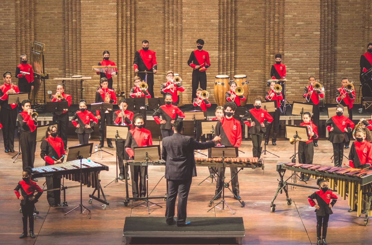 Na imagem uma orquestra composta por crianças e adolescentes se apresentam no palco do Auditório Claudio Santoro. Frente ao grupo está o mestra que rege a orquestra.
