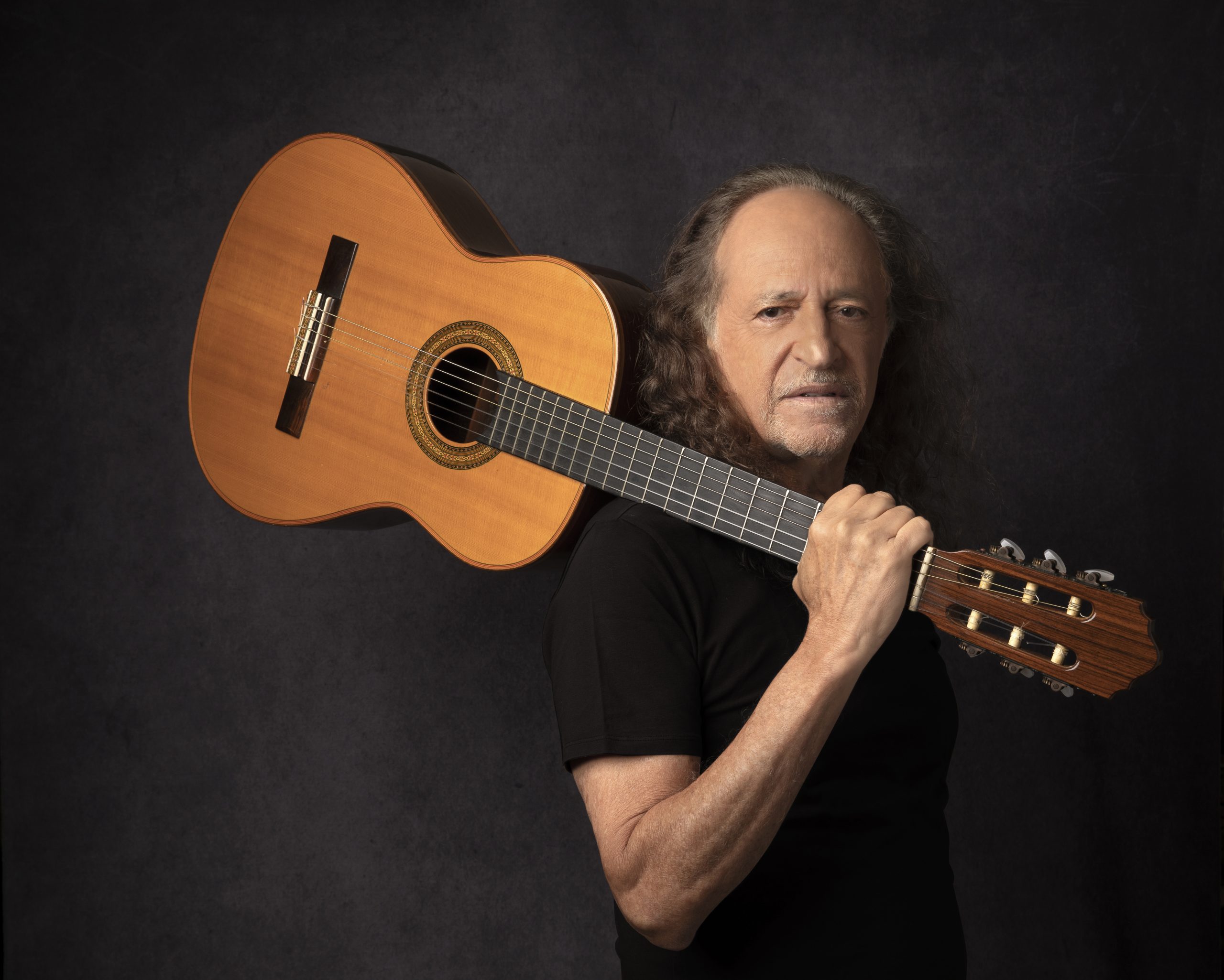 Foto do artista Alceu Valença segurando um violão apoiado no ombro. Ele está de camiseta preta e o fundo da arte também é preto.