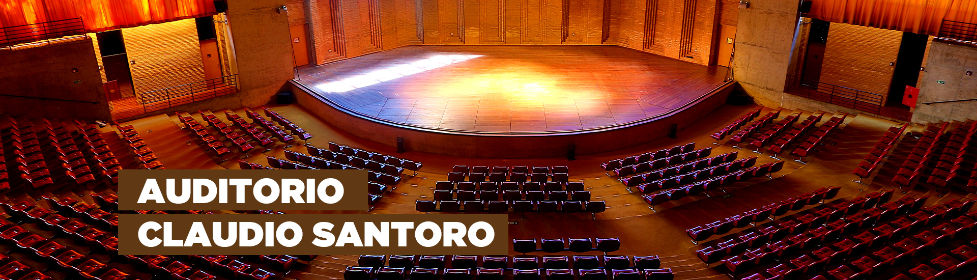A imagem mostra o Auditório Claudio Santoro que possui uma arquitetura que se assemelha a arena. Há 814 poltronas na cor vermelha, um palco com luzes e vidraças nas quais é possível observar a natureza. Na imagem está escrito "Auditorio Claudio Santoro"
