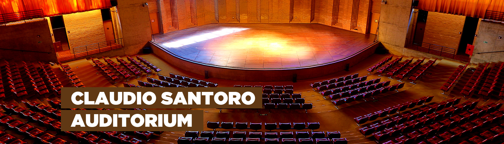 A imagem mostra o Auditório Claudio Santoro que possui uma arquitetura que se assemelha a arena. Há 814 poltronas na cor vermelha, um palco com luzes e vidraças nas quais é possível observar a natureza. Na imagem está escrito "Claudio Santoro Auditorium"