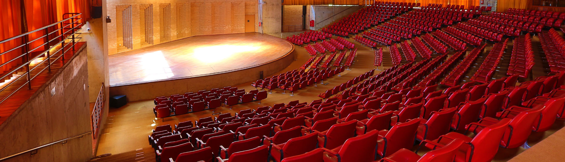 Na imagem há o palco do Auditório Claudio Santoro em primeiro plano e em segundo plano poltronas vermelhas divididas em cinco setores. Acima há estrutura arquitetônica do Auditório com recortes da laje. E aos fundos do espaço, há cortinas laranjas.