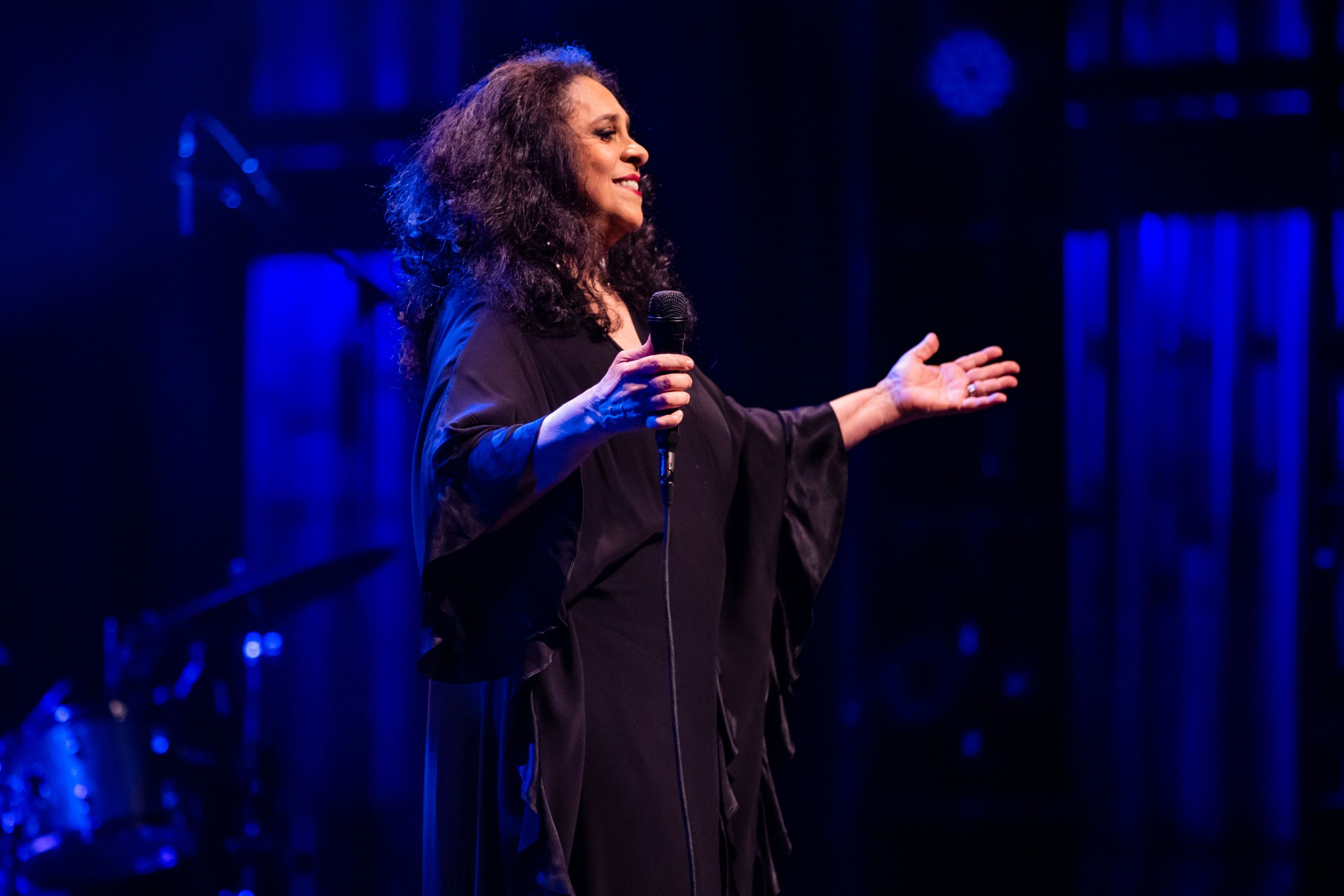 A imagem mostra a cantora Gal Costa que sorri com os braços abertos. Ela usa um vestido longo preto. O fundo da imagem possui tons pretos e azuis.