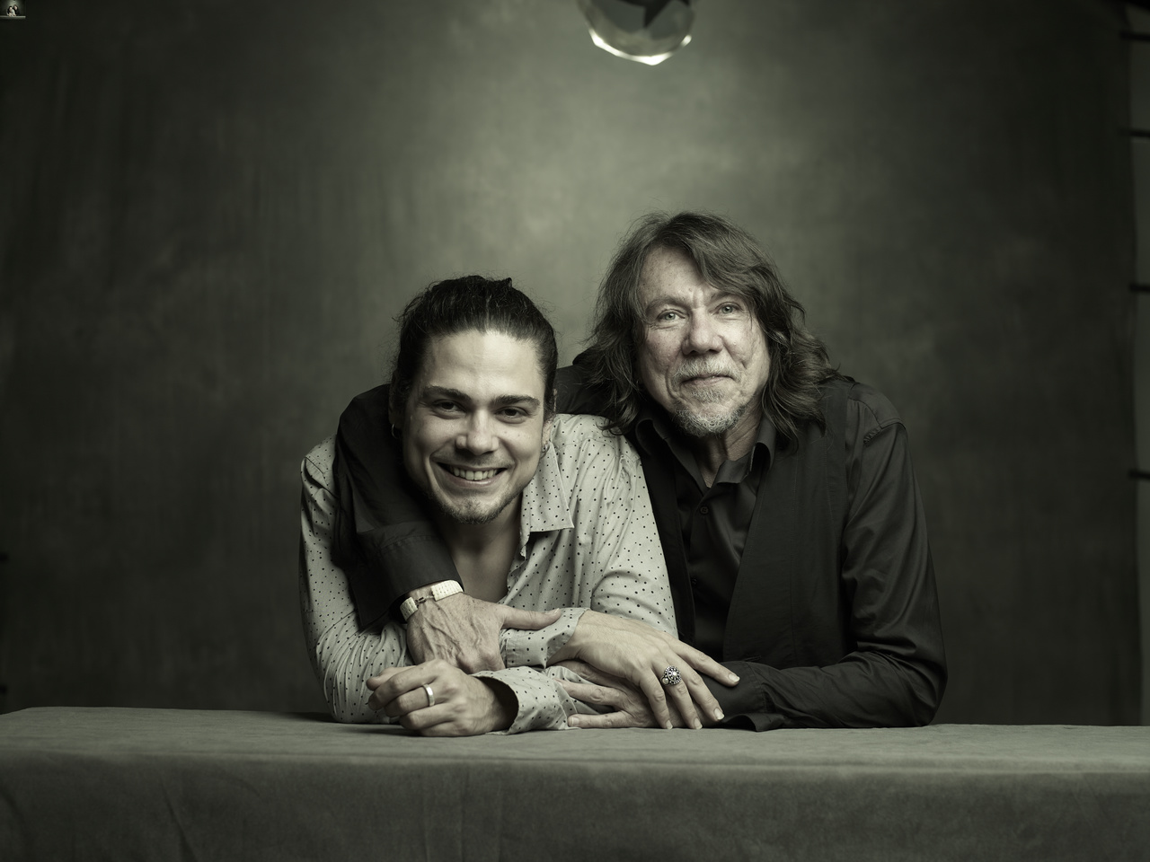 Foto dos artistas Bruno e Lenine abraçados. Bruno veste uma camisa preta e Lenine uma camisa cinza chumbo. Ambos estão sorrindo. O fundo da foto apresenta um degradê do preto para o bege, com tons bem escuros.