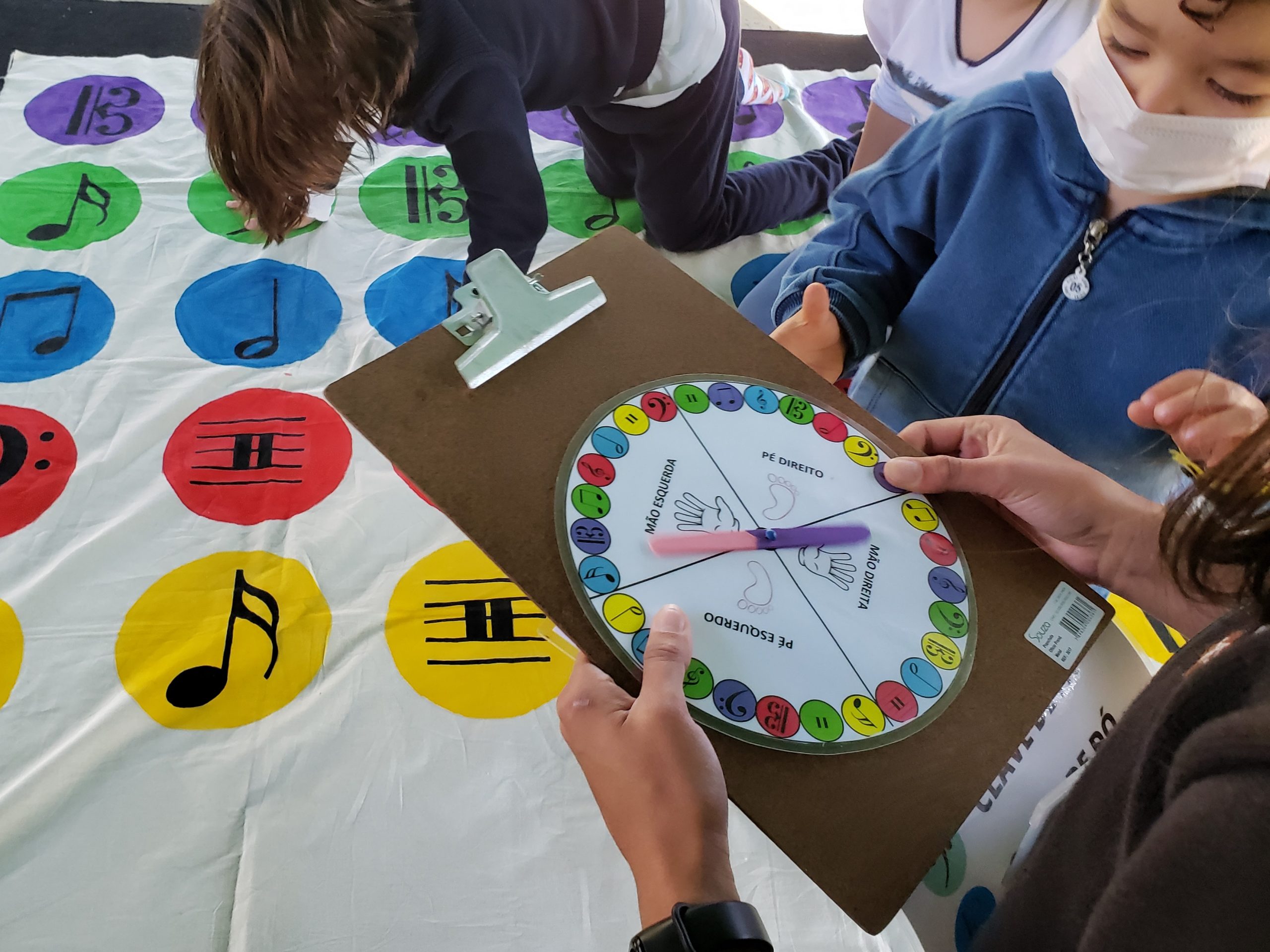 Na imagem há uma mão segurando uma prancheta com um disco giratório do jogo Twister Musical. No fundo da imagem há uma tapete do jogo com notas musicais nas cores roxa, verde, azul, vermelho e amarelo.