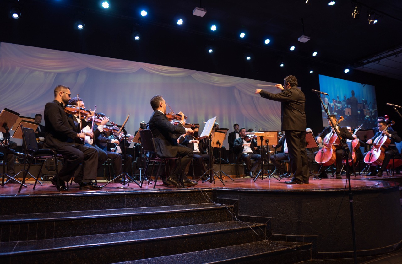 Na imagem a Orquestra Sinfônica Municipal de Barretos se apresenta num palco, no qual há um telão ao fundo com projeção de cortinas. Os músicos estão tocando diferentes instrumentos como violino e violoncelo e, seguem a orientação do mastro que está posicionado no meio do palco.