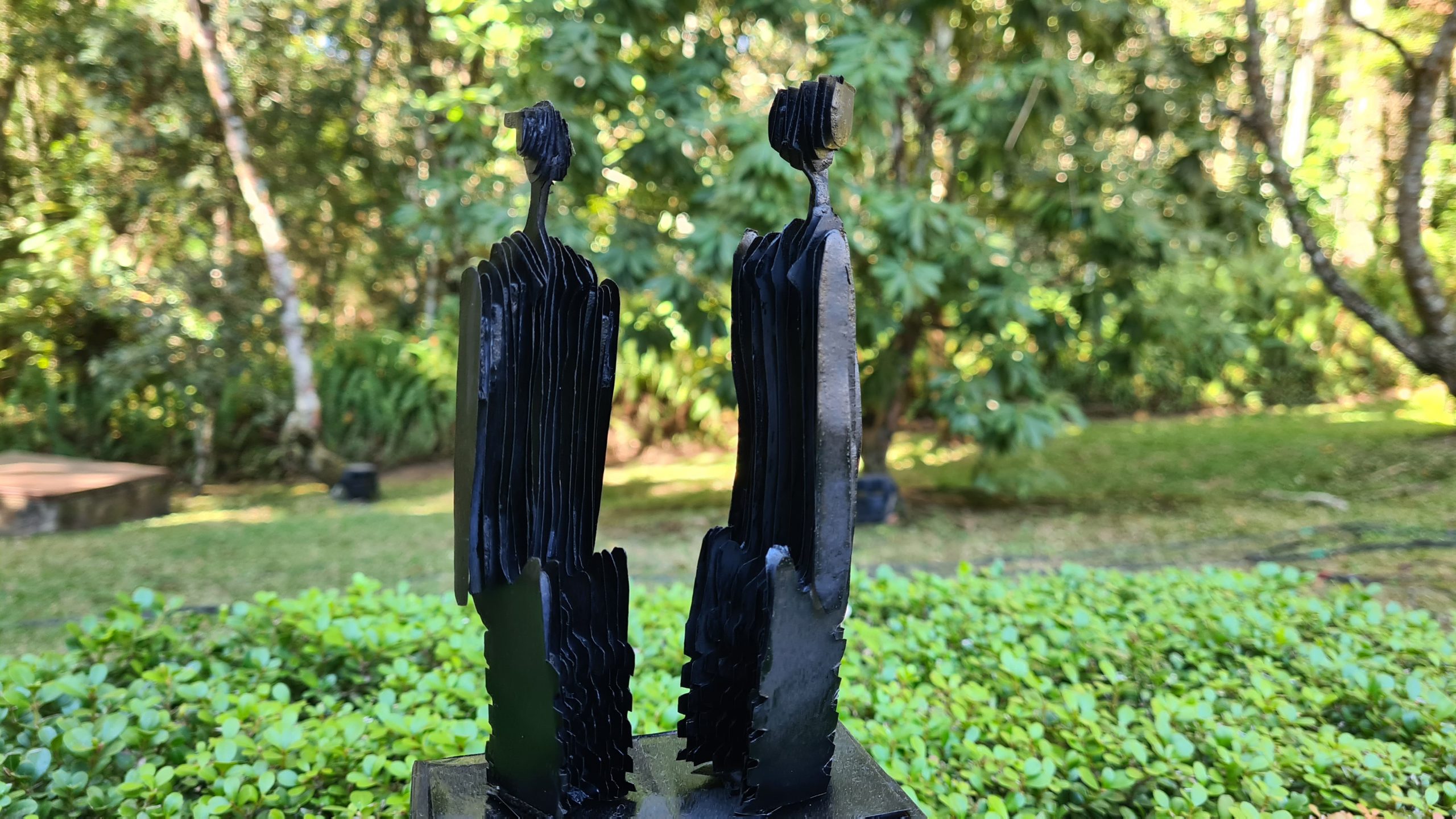 Uma escultura pequena feita a partir de recortes de papel na cor preta está em um espaço a céu aberto com mata nativa ao redor.