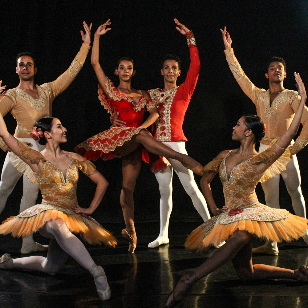 A imagem mostra o show da São Paulo Companhia de Dança, na qual uma bailarina com vestido vermelho e dourado ao lado de um bailarino vestindo uma blusa das mesmas cores, exceto pela calça, que é branca. Ambos estão curvados em um movimento de dança. Ao redor há duas bailarinas e dois bailarinos utilizam roupas em tons marrons.